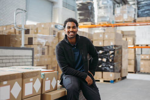 Fröhlicher Logistikmitarbeiter, der in die Kamera lächelt, während er auf einem Stapel von Paketkisten in einem Fulfillment-Center sitzt. Fröhlicher junger Mann, der in einem großen Auslieferungslager arbeitet. - JLPPF00456