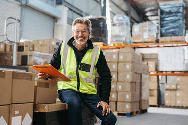 Happy senior man smiling at the camera while holding a clipboard in a distribution warehouse. Cheerful logistics manager wearing a reflective jacket in a large fulfillment centre. - JLPPF00457