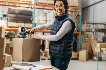 Happy warehouse worker sticking scotch tape on a cardboard box in a modern distribution centre. Mature woman smiling while preparing a package for shipment in a large fulfillment centre. - JLPPF00460
