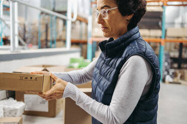 Mature warehouse employee packing cardboard boxes in a distribution centre. Female warehouse worker preparing packages for shipment in a large fulfilment centre. - JLPPF00461