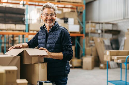 Älterer Mann, der in die Kamera lächelt, während er in einem Auslieferungslager Kartons verpackt. Glücklicher Logistikmitarbeiter, der in einem großen Fulfillment-Center Waren für den Versand vorbereitet. - JLPPF00466