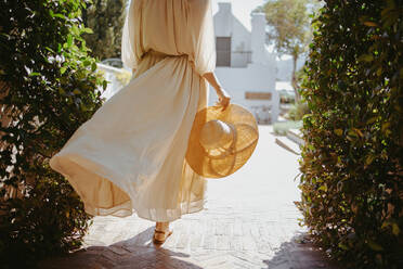 Anonyme Touristin, die in einem fließenden, eleganten Kleid spazieren geht. Rückansicht einer unerkennbaren jungen Frau, die alleine vor einem luxuriösen Urlaubsort spazieren geht. Junge Frau, die im Sommer alleine Urlaub macht. - JLPPF00491