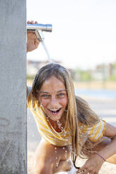 Fröhliches Teenager-Mädchen, das lächelnd in die Kamera schaut, während es unter einem Wasserstrahl auf einer Böschung steht, vor einem unscharfen Hintergrund an einem Sommertag - ADSF38766