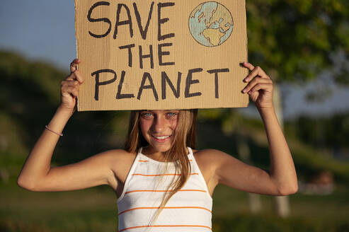 Smiling teenage girl holding cardboard poster with Save The Planet inscription standing looking at camera on street park on summer day - ADSF38767