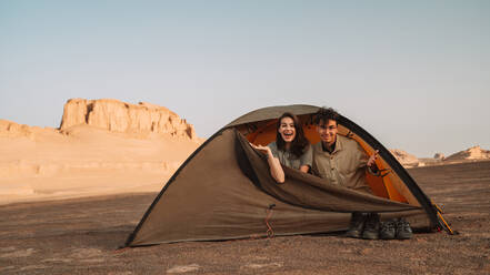 Happy couple sitting in tent during trip in Lut desert with rough rocky cliffs on sunny summer day in Iran - ADSF38803