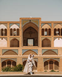 Full body of female in authentic outfit standing on street near historic mosque with ornamental elements on sunny summer day in Iran - ADSF38806