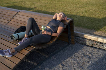 Happy woman holding water bottle relaxing on wooden bench - VPIF07437