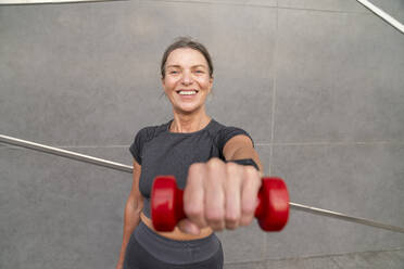 Happy woman showing red dumbbell in front of wall - VPIF07454