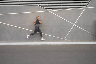 Mature woman jogging on footpath by gray wall - VPIF07462