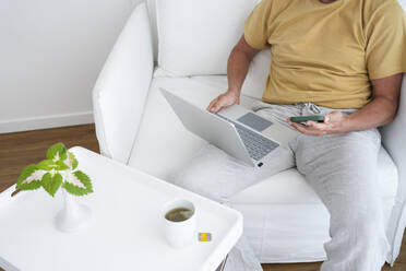 Freelancer with laptop using smart phone sitting in armchair at home - SVKF00537