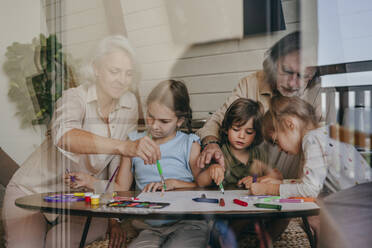 Großeltern mit Kindern, die zu Hause malen, gesehen durch das Glas eines Fensters - YTF00122
