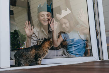 Glückliche Großmutter und Enkelin, die eine Krone tragen und eine Katze durch ein Glasfenster betrachten - YTF00132