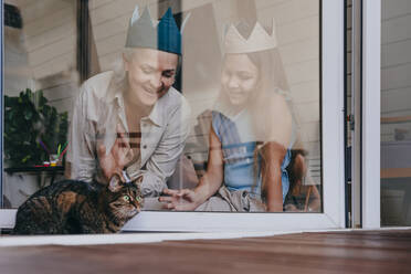 Glückliche Enkelin und Großmutter mit Krone, die durch ein Glasfenster eine Katze betrachten - YTF00133