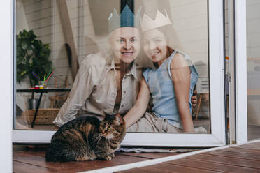 Glückliche Großmutter und Enkelin mit Krone sitzen zusammen hinter einem Glasfenster zu Hause - YTF00134