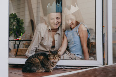 Lächelnde Enkelin und Großmutter mit Krone, die durch ein Glasfenster eine Katze betrachten - YTF00135