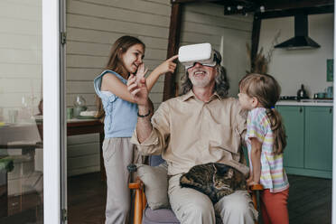 Lächelnder Großvater mit VR-Brille gestikuliert bei den Enkelinnen zu Hause - YTF00138
