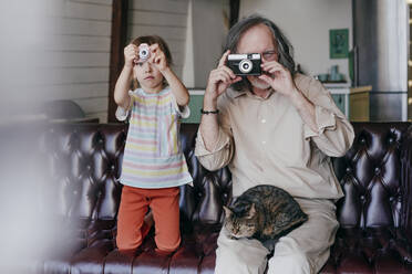 Großvater mit alter Kamera und Enkelin mit Spielzeugkamera beim Fotografieren auf dem Sofa - YTF00158