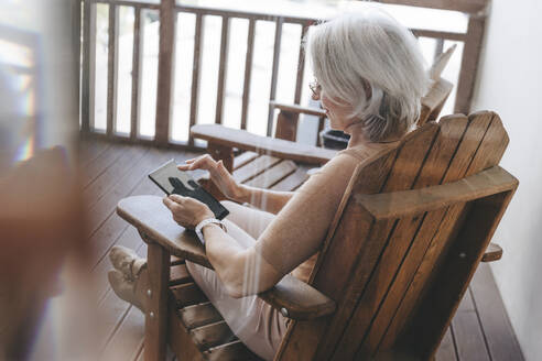 Ältere Frau sitzt auf einem Stuhl und benutzt einen Tablet-PC auf der Terrasse zu Hause - YTF00163