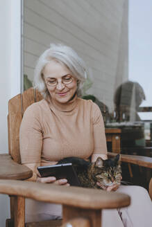 Lächelnde reife Frau mit Tablet-PC und Katze auf dem Schoß auf der heimischen Terrasse - YTF00164