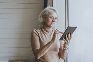 Lächelnde Frau, die einen Tablet-PC benutzt und zu Hause am Fenster steht - YTF00166