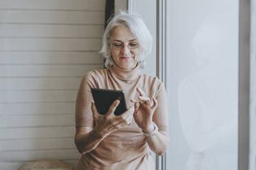 Smiling mature woman standing near window and using tablet PC - YTF00167