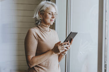 Mature woman with gray hair using tablet PC by window at home - YTF00168