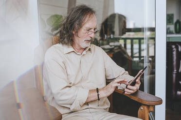 Älterer Mann mit Tablet-PC auf einem Stuhl zu Hause sitzend - YTF00170