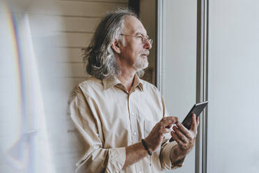 Nachdenklicher älterer Mann mit Tablet-PC am Fenster zu Hause - YTF00173