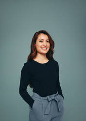 Happy young woman smiling at the camera in a studio. Independent young woman standing with her hands in her pockets against a studio background. Cheerful young woman embracing girl power. - JLPPF00506