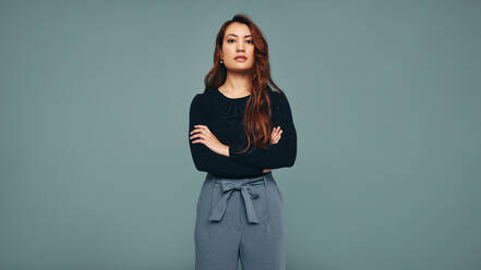 Independent young woman standing in a studio. Confident young woman looking at the camera while standing with her arms crossed against a studio background. Strong young woman embracing girl power. - JLPPF00509