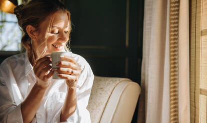 Lächelnde junge Frau, die eine Tasse Kaffee in einem Hotel genießt. Attraktive junge Frau, die sich auf einem Stuhl in ihrem Hotelzimmer entspannt. Glückliche junge Frau, die ihren Sommerurlaub in einem Luxushotel genießt. - JLPPF00650