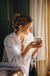 Happy young woman enjoying a cup of coffee in a hotel. Attractive young woman relaxing on a chair in a white shirt. Young woman enjoying her summer vacation in a luxury hotel. - JLPPF00651