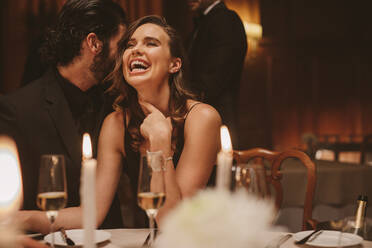 Ein Mann flüstert einer lächelnden Frau auf einer Dinnerparty etwas Lustiges ins Ohr. Ein fröhliches Paar genießt ein Gala-Dinner. - JLPPF00664