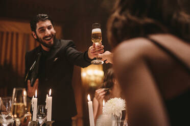 Gutaussehender junger Mann reicht einer Freundin, die am Esstisch sitzt, ein Glas Champagner. Freunde trinken bei einer Dinnerparty. - JLPPF00690