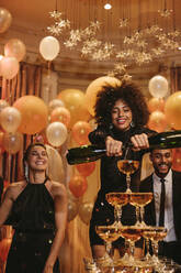 Afrikanische junge Frau Füllung von Champagner Pyramide mit Freunden im Hintergrund an Silvester. Multi-ethnischen geselligen Menschen genießen bei einer Gala-Nacht-Party. - JLPPF00711
