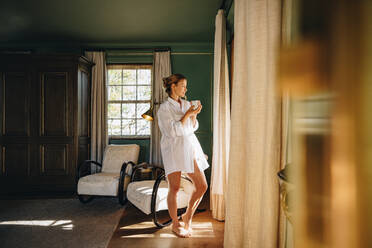 Vacationing in a luxury hotel. Attractive young woman looking outside the window while standing alone in a hotel room. Woman holding a cup of coffee while wearing a white shirt. - JLPPF00758