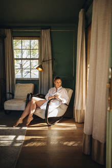 Sommerurlaub in einem Luxushotelzimmer. Attraktive junge Frau, die aus dem Fenster schaut, während sie sich auf einem Stuhl in einem weißen Hemd entspannt. Junge Frau, die ihren Urlaub in einem Luxushotel genießt. - JLPPF00761