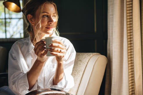 Junge Frau schaut aus ihrem Hotelfenster. Attraktive junge Frau mit einer Tasse Kaffee in der Hand, während sie sich auf einem Stuhl entspannt. Gelassene junge Frau, die ihren Sommerurlaub in einem Luxushotel genießt. - JLPPF00763