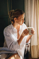 Unbeschwerte Frau trinkt Kaffee in ihrem Hotelzimmer. Attraktive junge Frau mit einer Tasse Kaffee in der Hand, während sie sich auf einem Stuhl entspannt. Junge Frau, die ihren Sommerurlaub in einem Luxushotel genießt. - JLPPF00765