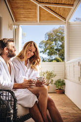 Relaxing on the hotel balcony. Happy couple smiling cheerfully while having coffee together in the morning. Married couple enjoying a romantic weekend getaway at a luxury hotel. - JLPPF00784