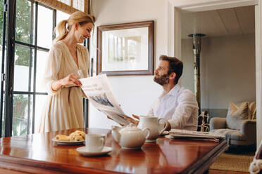 Smiling wife taking away her husband's newspaper inside a hotel. Happy woman standing in front of her husband at a dining table. Romantic couple enjoying a lovely morning during their vacation. - JLPPF00812