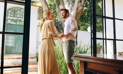Middle aged couple laughing together while standing outside a hotel entrance. Happy couple enjoying a summer morning together. Married couple spending a weekend getaway at a luxury hotel. - JLPPF00821