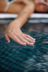 Weibliche Hand, die die Wasseroberfläche in einem Schwimmbad berührt. Unerkennbare Touristin, die ihre Hand über das Wasser streckt, während sie am Pool liegt. Frau, die in einem Luxus-Spa-Resort Urlaub macht. - JLPPF00852