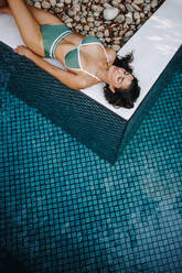 Draufsicht auf eine heitere junge Frau, die sich am Pool entspannt. Attraktive junge Touristin, die im Bikini am Pool liegt. Frau, die ihren Sommerurlaub in einem Luxusferienort genießt. - JLPPF00854