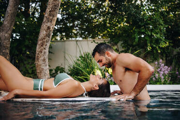 Flitterwöchner flirten an einem luxuriösen Swimmingpool. Ein glückliches junges Paar lächelt sich an, während es sich am Pool entspannt. Ein junges Paar genießt seinen Flitterwochenurlaub in einem luxuriösen Wellness-Resort. - JLPPF00856