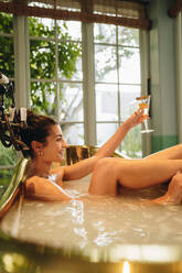 Smiling young woman toasting with champagne in a bathtub. Attractive young woman raising a glass of champagne while relaxing in a bathtub. Young woman enjoying a vacation at a luxury hotel. - JLPPF00904