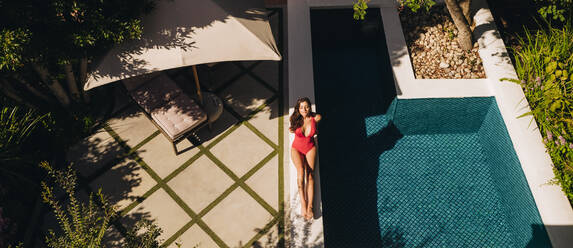 Hohe Winkelansicht einer jungen Frau, die sich in einem roten Badeanzug am Rande eines Swimmingpools sonnt. Attraktive junge Frau, die sich außerhalb eines Luxus-Spa-Resorts entspannt. Junge Frau, die ihren Sommerurlaub genießt. - JLPPF00912