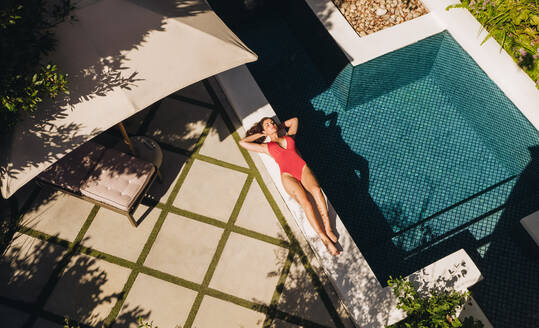 Relaxing by the poolside. High angle view of an attractive young woman sunbathing on the edge of a swimming pool in a red swimsuit. Young woman enjoying a weekend getaway at a luxury spa resort. - JLPPF00915