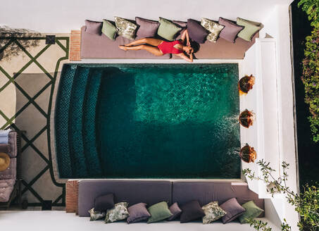 Luftaufnahme einer attraktiven jungen Frau, die sich in einem roten Badeanzug sonnt. Junge Touristin, die neben einem Swimmingpool im Freien liegt. Frau, die ihren Sommerurlaub in einem Luxus-Spa-Resort genießt. - JLPPF00925