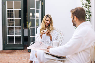 Carefree woman chatting with her husband over tea. Happy woman smiling while sitting outdoors with her husband in the morning. Married couple enjoying a getaway at a holiday resort. - JLPPF00933